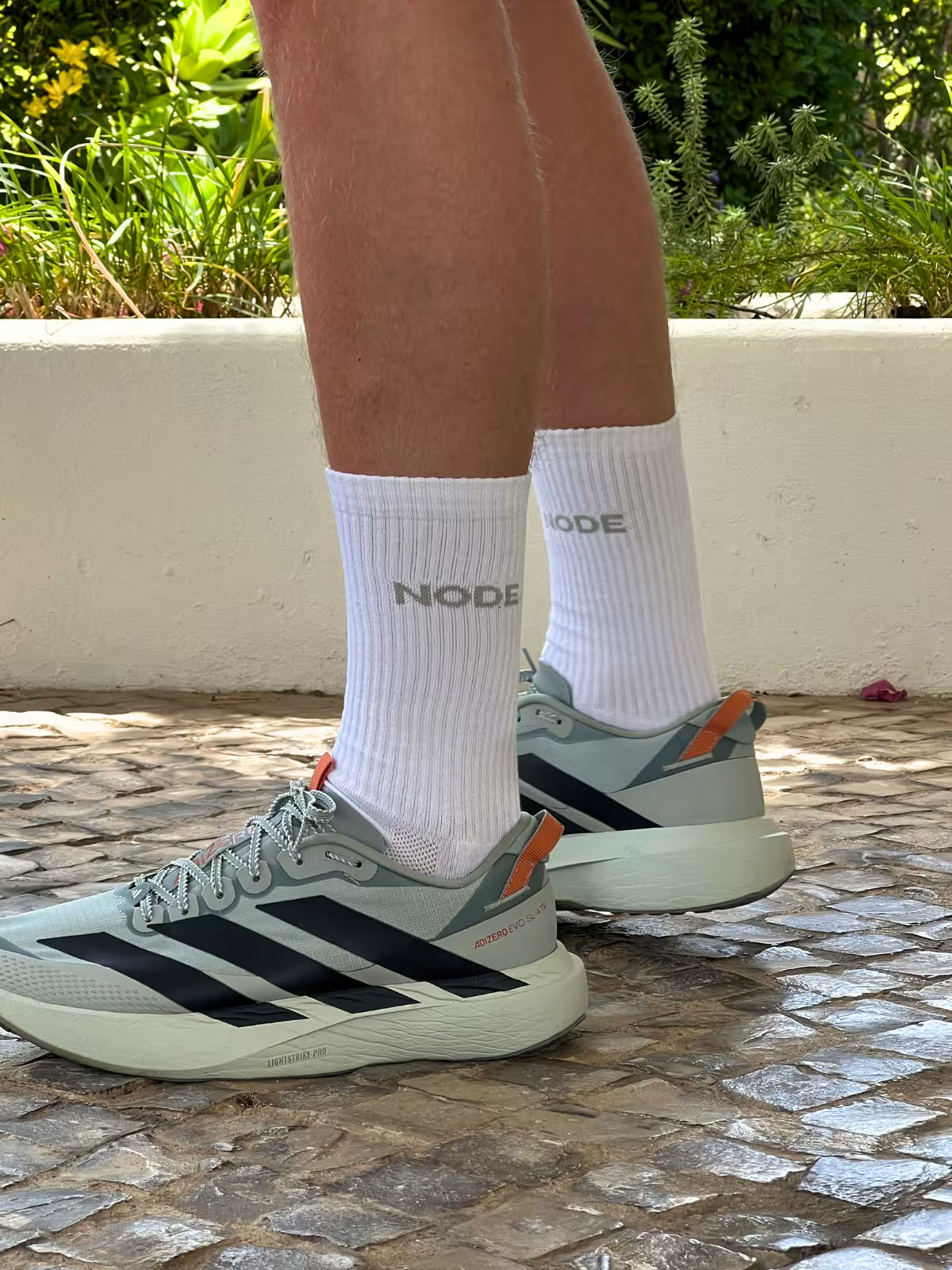 Person wearing white socks with 'NODE' branding and Adidas shoes on a stone pavement.
