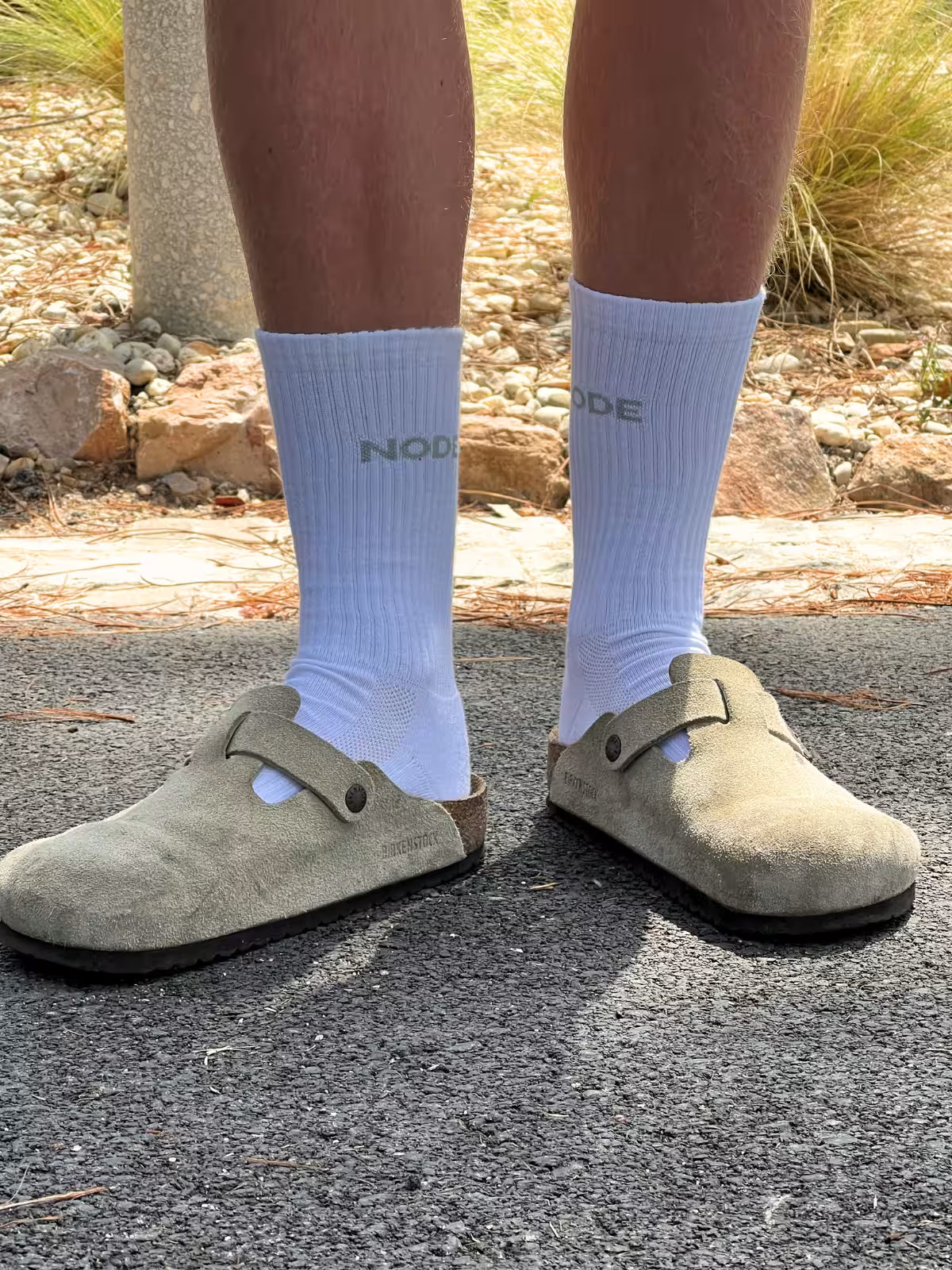 Person wearing light Taupe suede clog shoes and white socks with 'NODE' branding on a natural background.