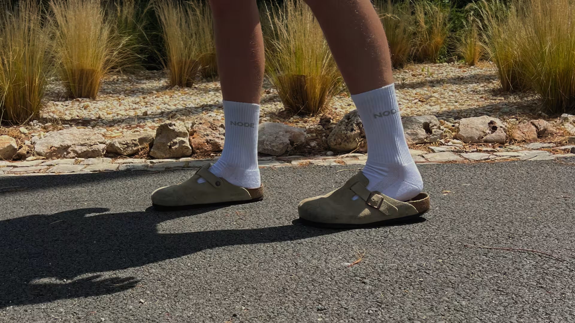 Model wearing white NODE socks and Taupe clog shoes on a paved surface with grass in the background